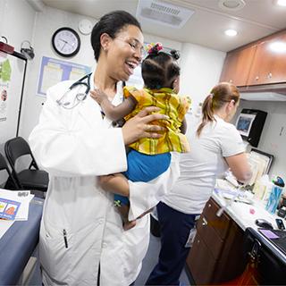 In an examination room, a smiing medical school student with a stethoscope around her neck holds a baby